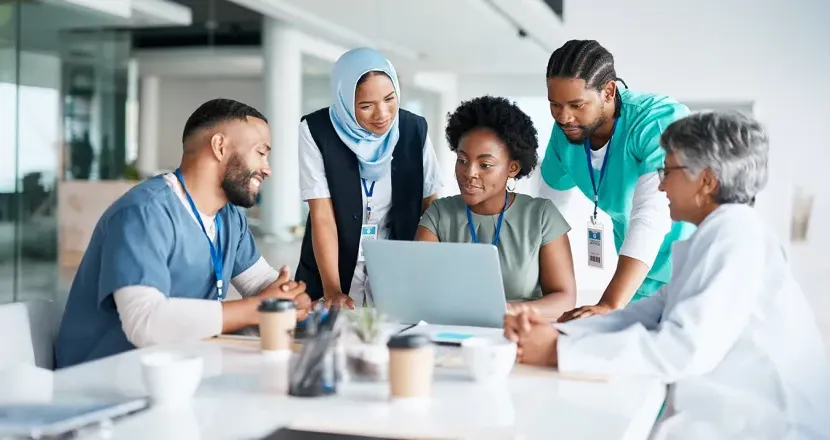 Diverse healthcare professionals collaborating around a laptop during a meeting, demonstrating teamwork and leadership in healthcare administration.